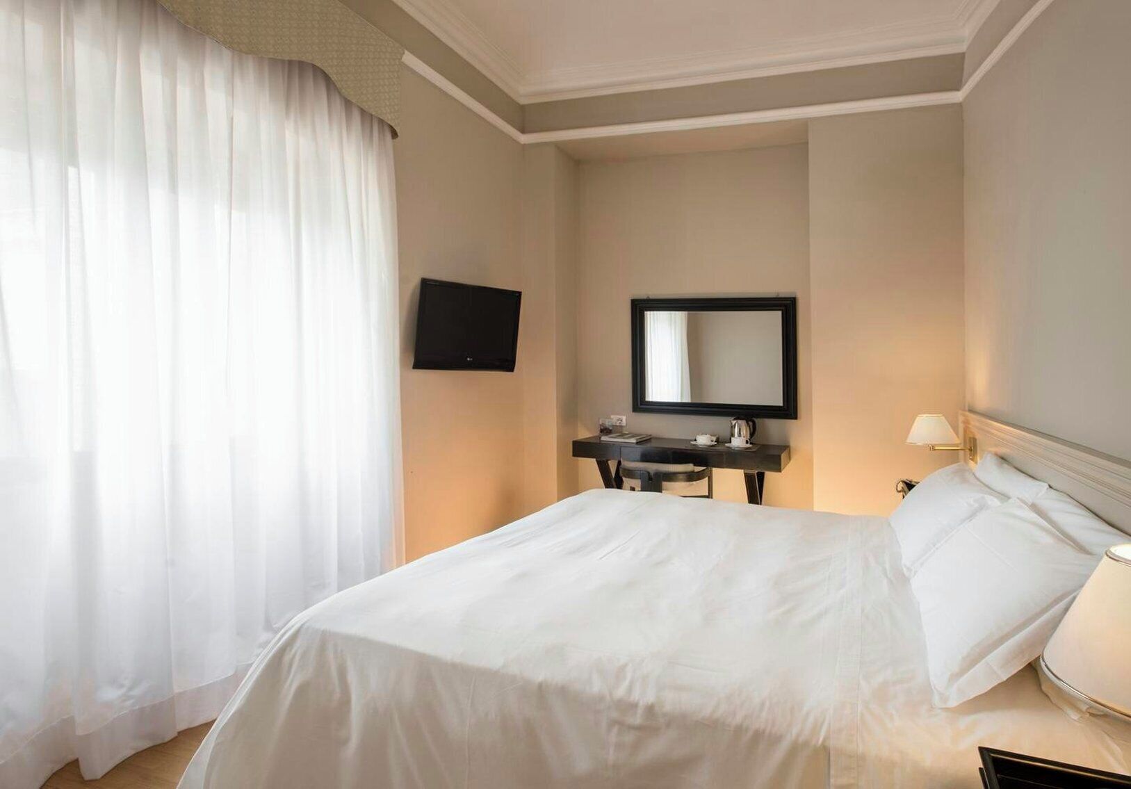 A hotel room with a bed, a desk with a flat-screen TV, a mirror, and a lamp.
