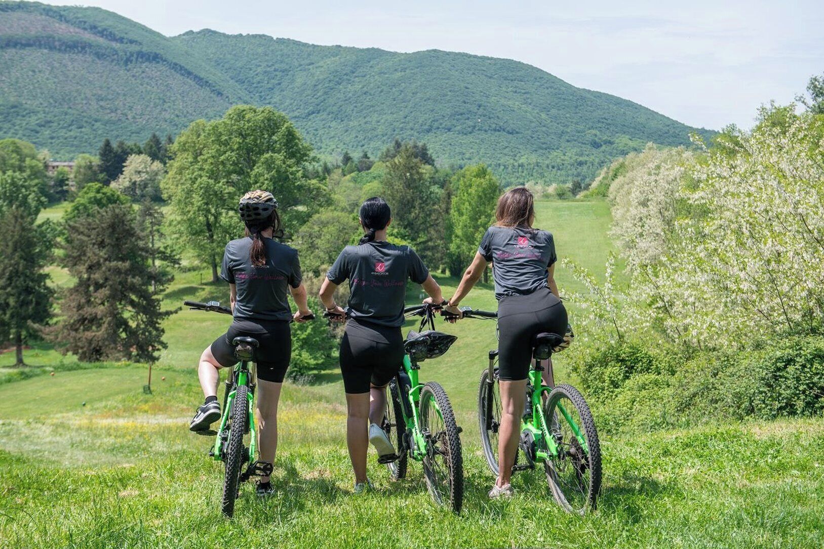 Trois personnes à vélo dans un champ herbeux, avec des montagnes en arrière-plan.