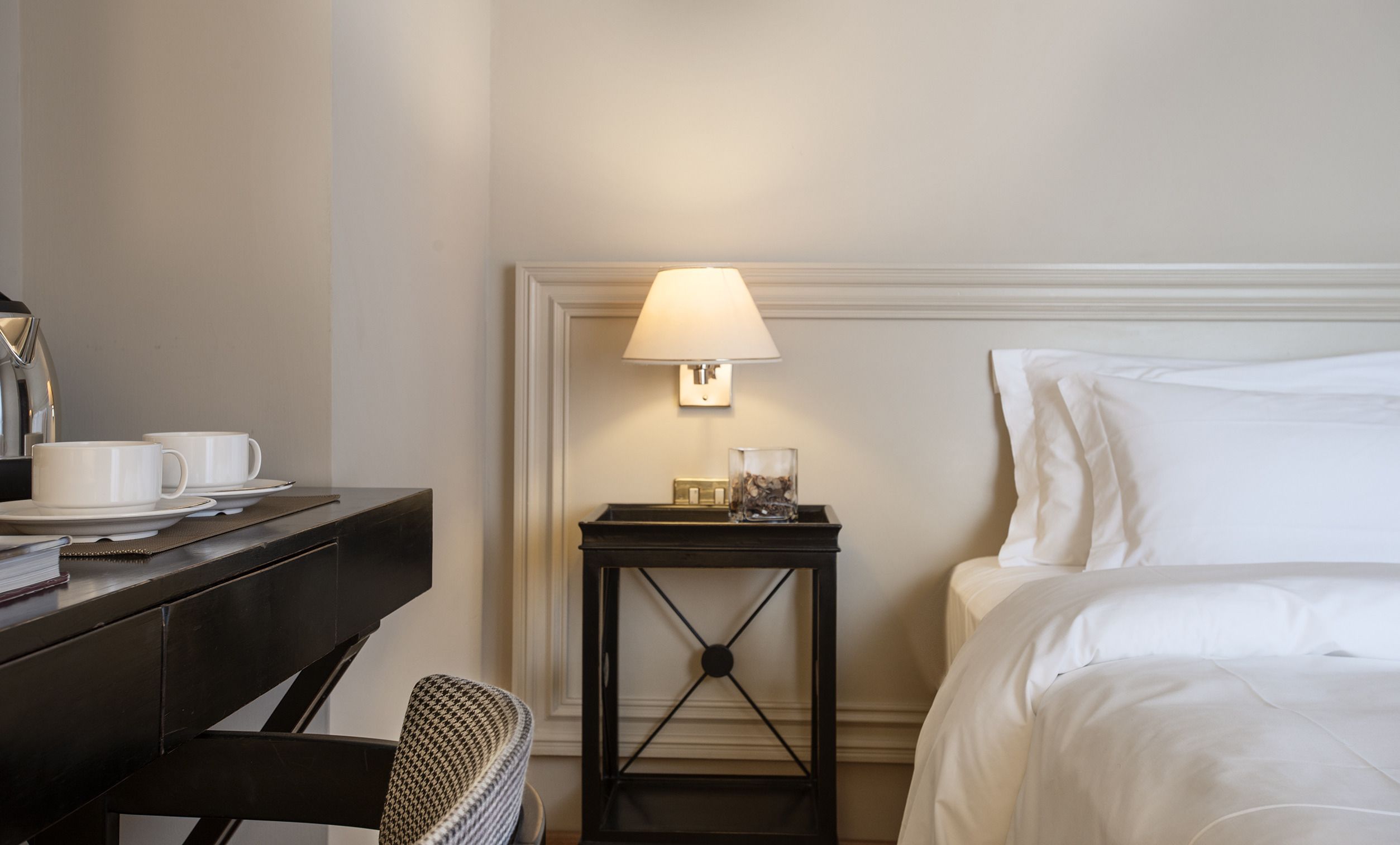 A hotel room with a bed, a desk with two white cups, a lamp, and a small table with a glass.