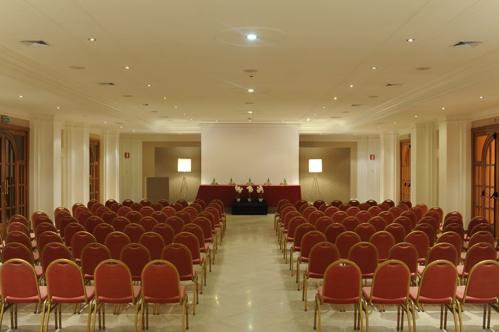 A conference room with rows of red chairs, a podium at the front, and a large screen.