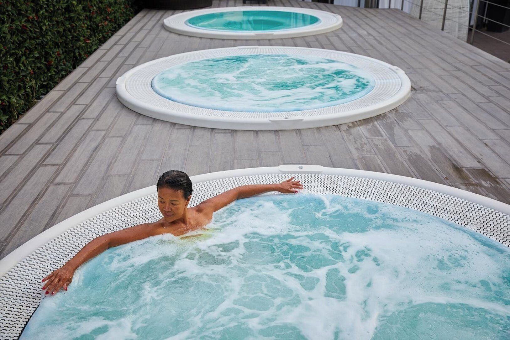 A person relaxing in a hot tub with a tiled deck and a hedge in the background.