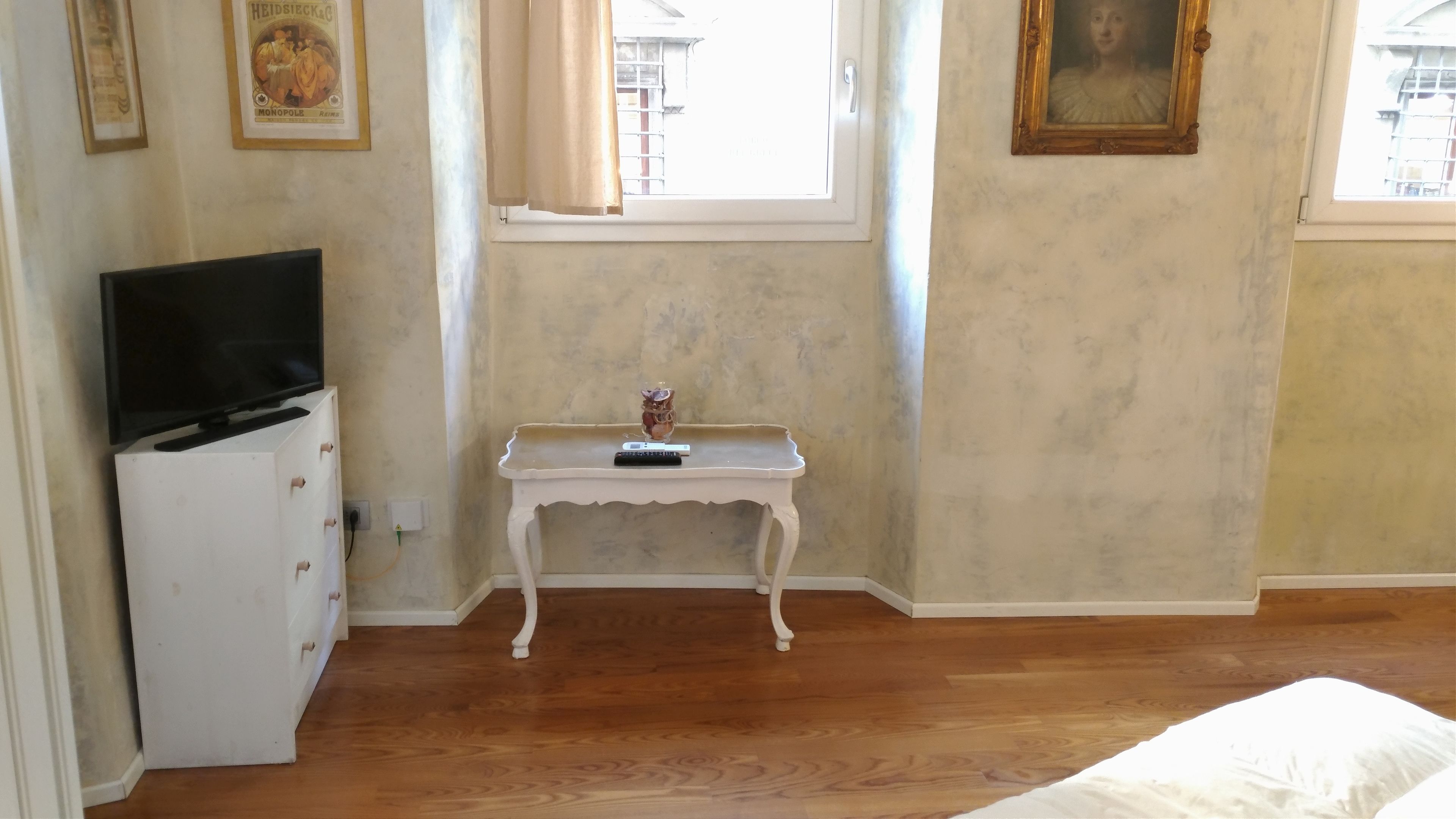 Ein Schlafzimmer mit einem Bett, einem Schminktisch, einem kleinen Tisch mit Fernbedienung, einem gerahmten Bild an der Wand und einem Fenster mit Vorhängen.