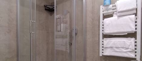 A glass-enclosed shower with a white towel on the floor, beige tiled walls, and a towel rack with multiple towels.