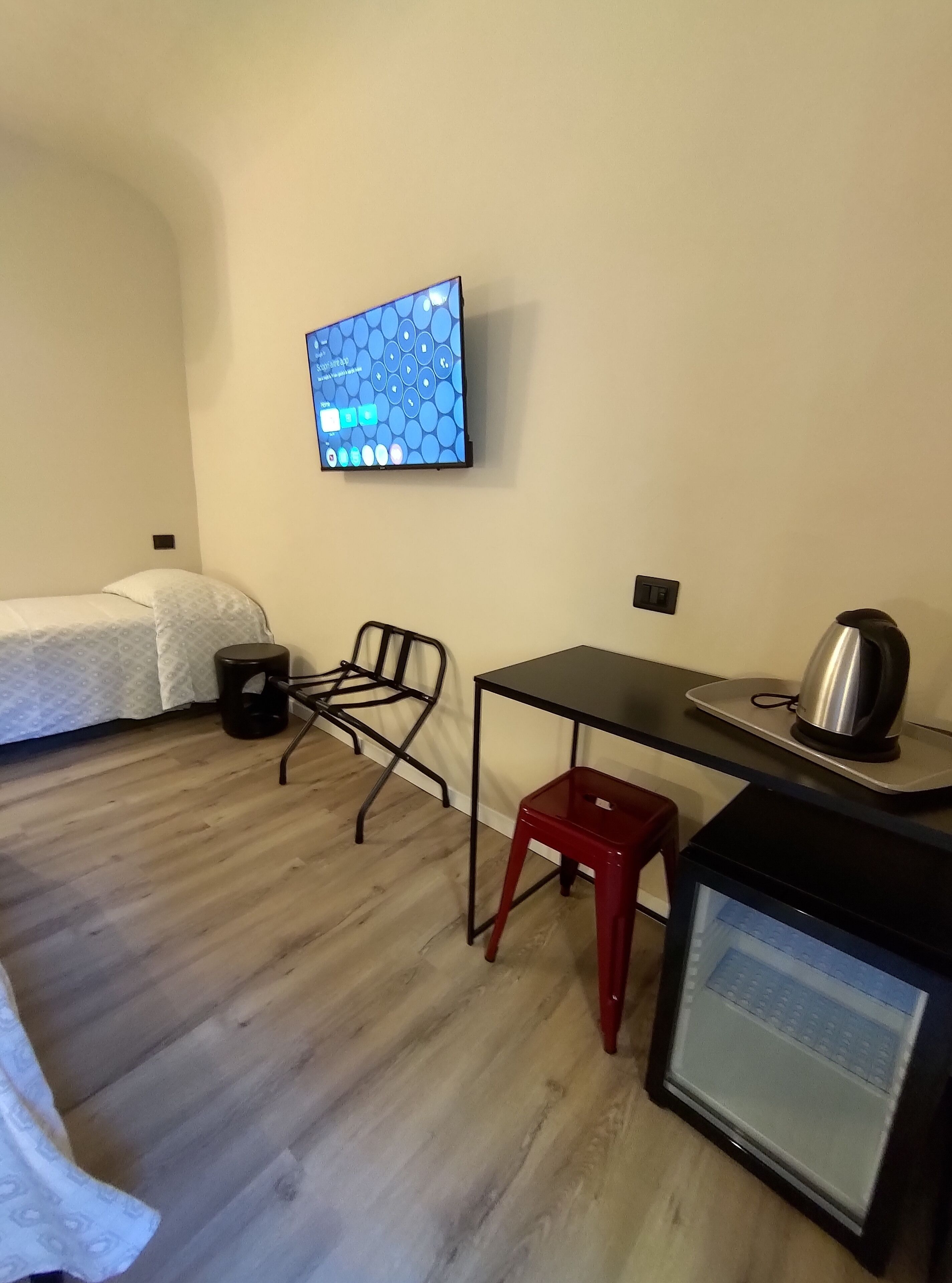 A small, simple room with a bed, a desk, a chair, a small stool, and a television.