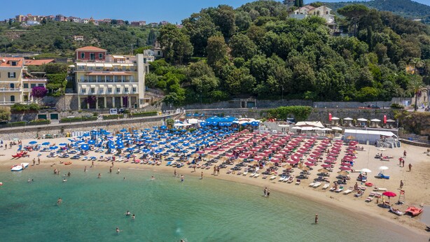 A beach with rows of sunbeds and umbrellas, a clear blue sky, and buildings in the background. - Hotel Venere Azzurra (Lerici)