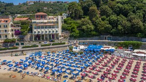 A beach with rows of blue and red umbrellas, a stone wall, and buildings in the background. - Hotel Venere Azzurra (Lerici)