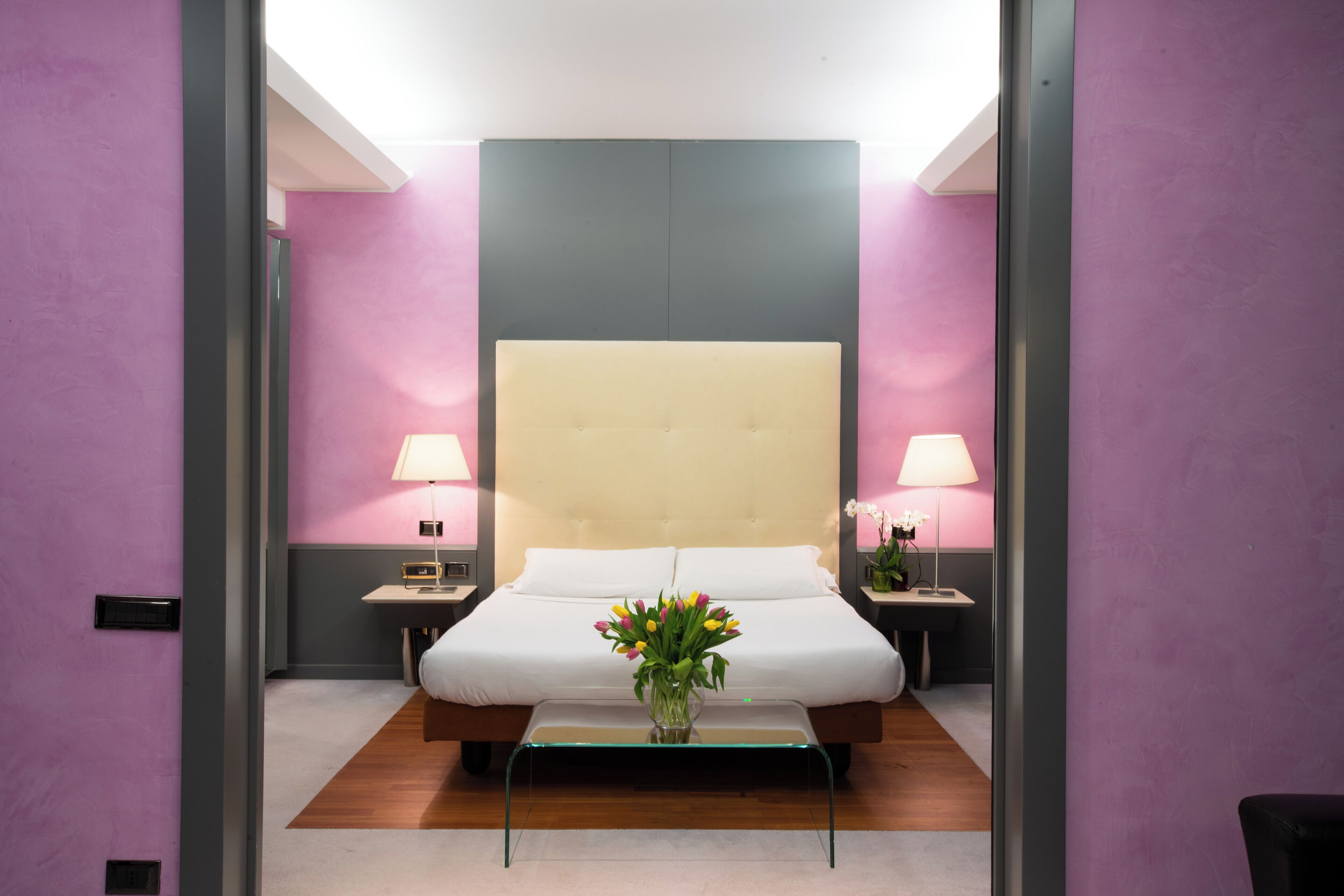 A modern hotel room with a large bed, bedside lamps, a small table with a vase of flowers, and a wooden floor.