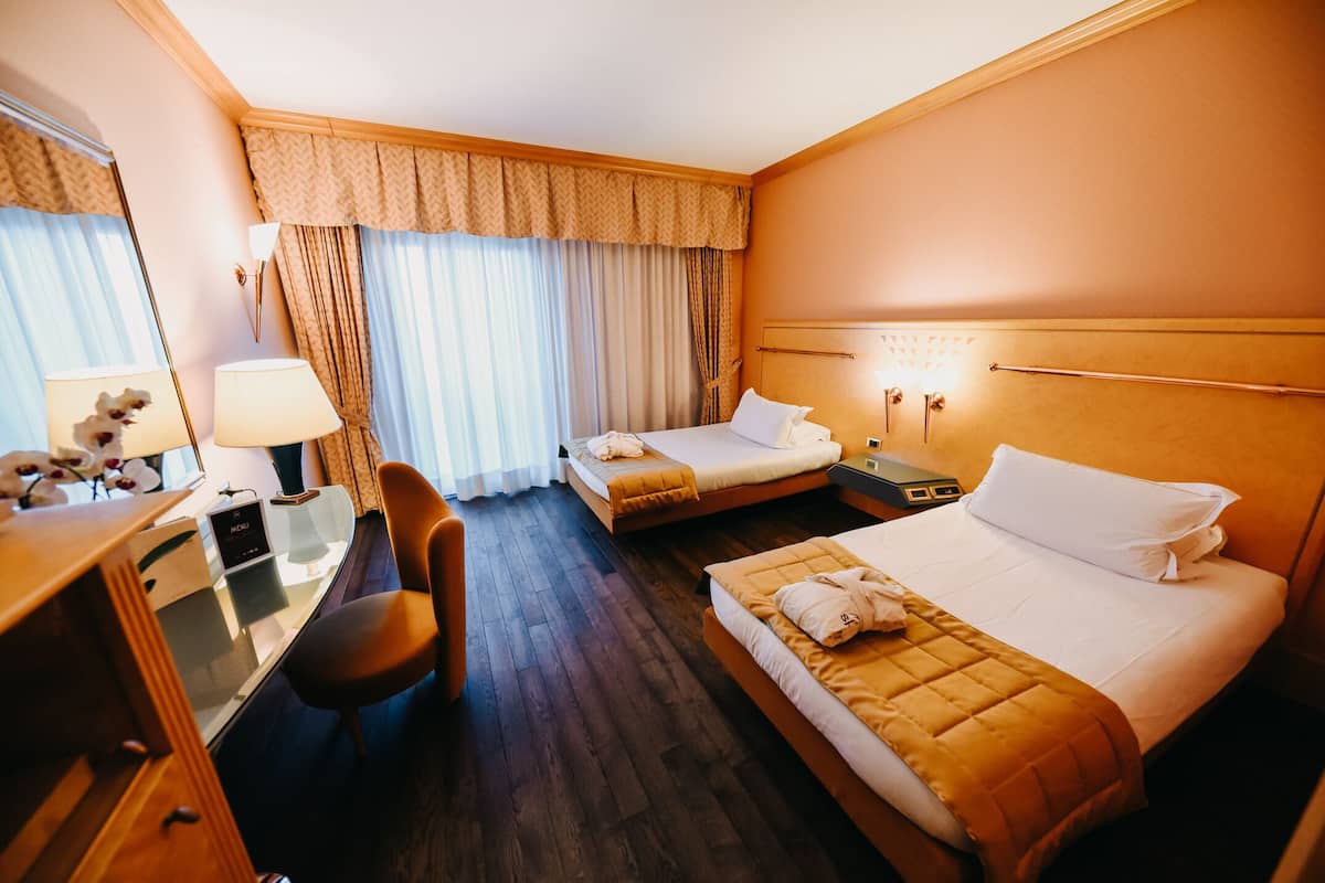 A hotel room with two beds, a desk, a chair, and a nightstand with a lamp.