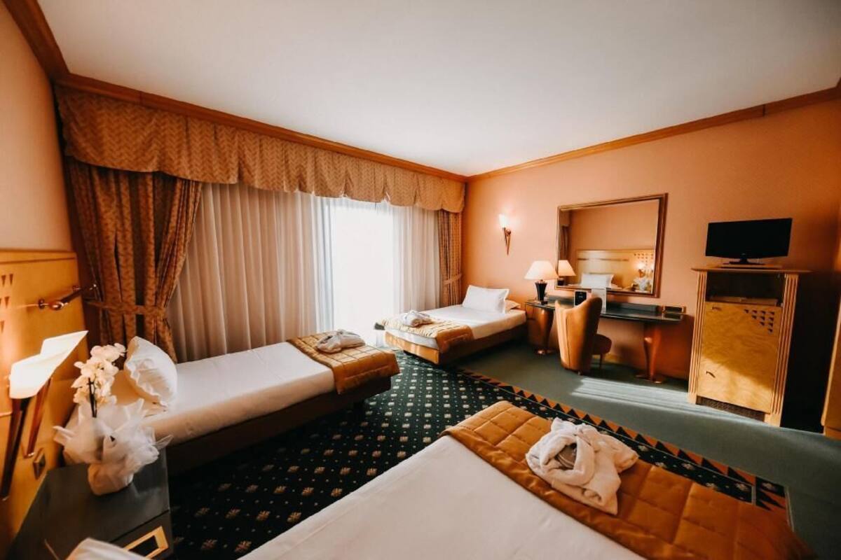 A hotel room with two beds, a desk, a chair, a television, and a large window with curtains.