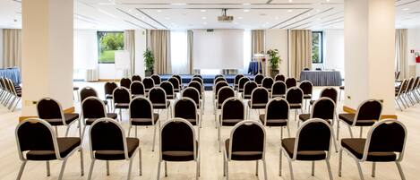 Un salón de conferencias con filas de sillas, una pantalla de proyección y una mesa con mantel azul.