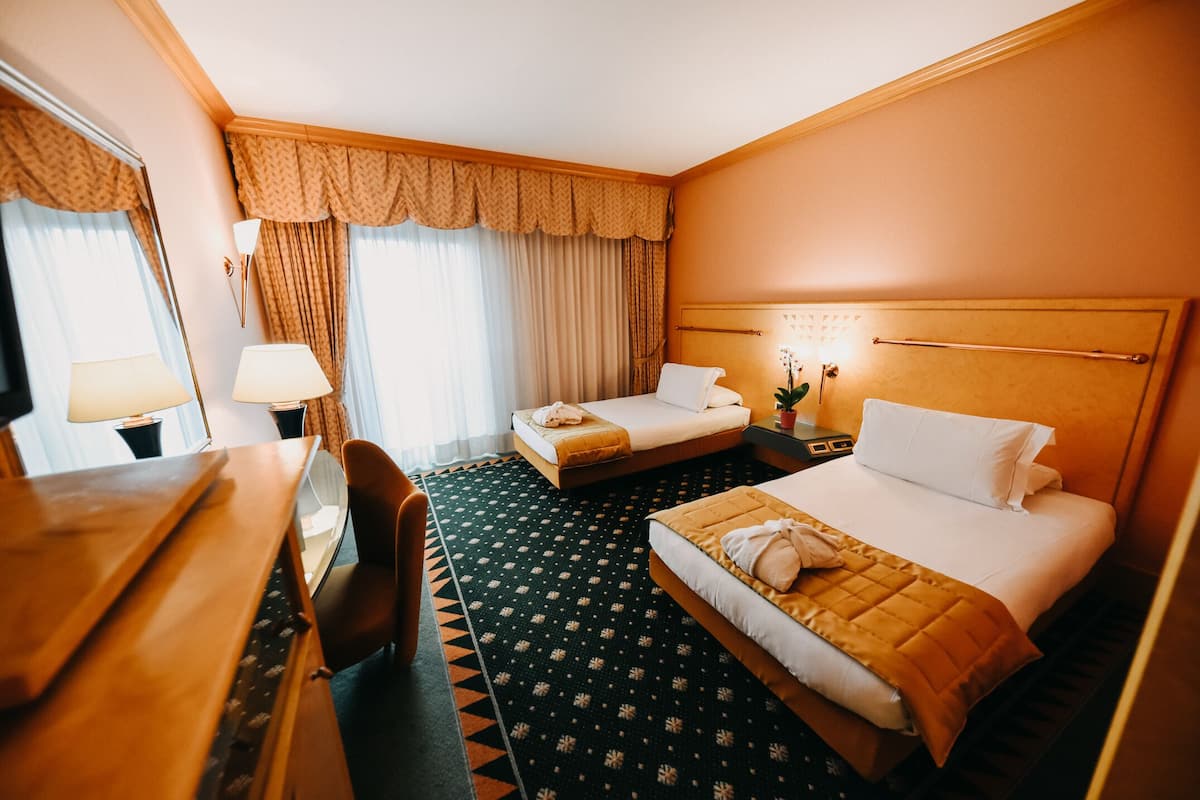 A hotel room with two beds, a desk, a chair, and a nightstand with a lamp.