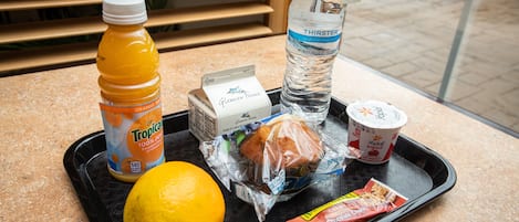 Una bandeja con una botella de jugo de naranja, una botella de agua, un limón, un sándwich, un vaso de yogurt y una bolsa de snacks.