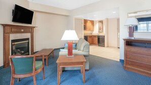 A hotel room with a TV mounted on the wall, a fireplace, a dining table with chairs, a sofa, and a kitchen area in the background.