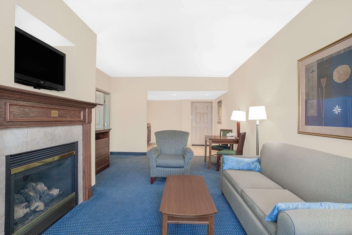 A hotel room with a fireplace, a television mounted on the wall, a striped armchair, a sofa, a small table, and a painting on the wall.