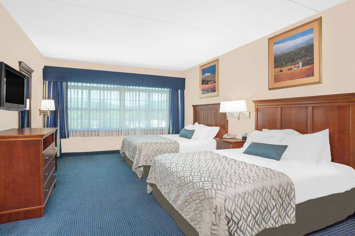 A hotel room with two beds, a TV, a dresser, and a large window with curtains.
