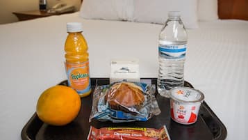 Bandeja con una botella de jugo de naranja Tropicana, una botella de agua, un paquete de trozos de chocolate, un pastel envuelto y un recipiente de yogur sobre una cama.