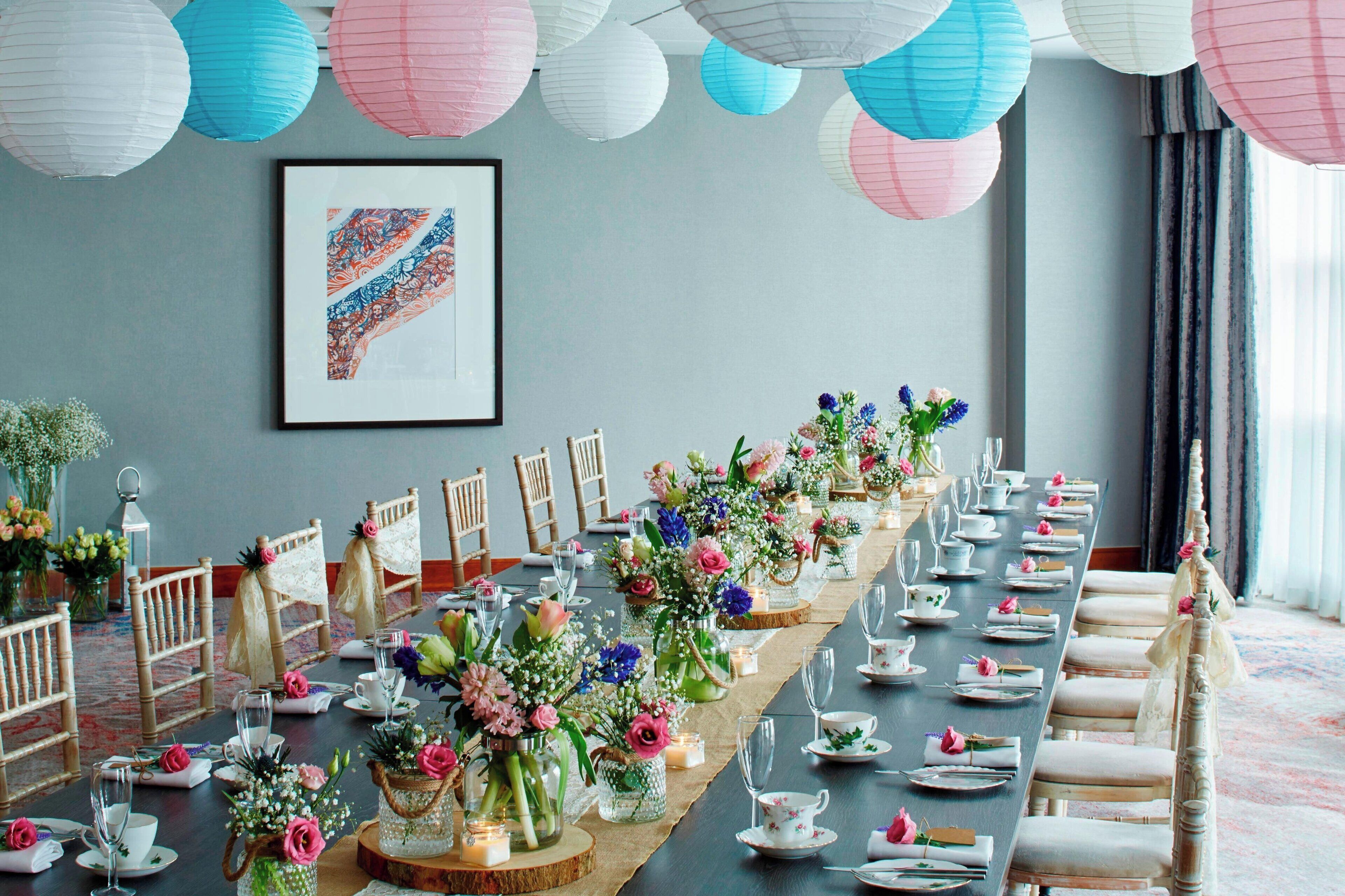 A long dining table set with floral centerpieces, white and pink paper lanterns, and a framed abstract artwork on the wall.