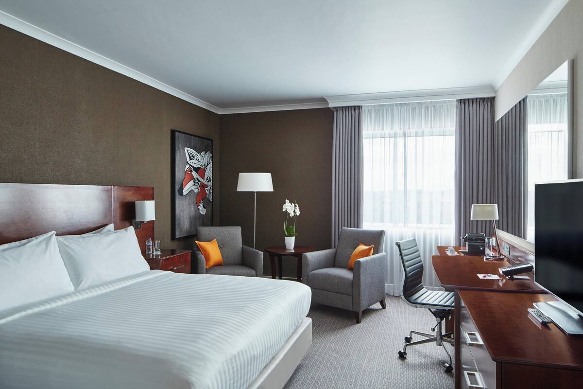 A hotel room with a large bed, a desk with a computer, a chair, a small table with a lamp, and a window with curtains.