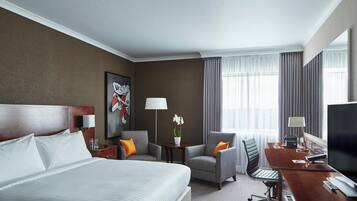 Ein Hotelzimmer mit einem großen Bett, einem Schreibtisch mit Computer, einem Sessel, einem kleinen Tisch mit Lampe und einem Fenster mit Vorhängen.