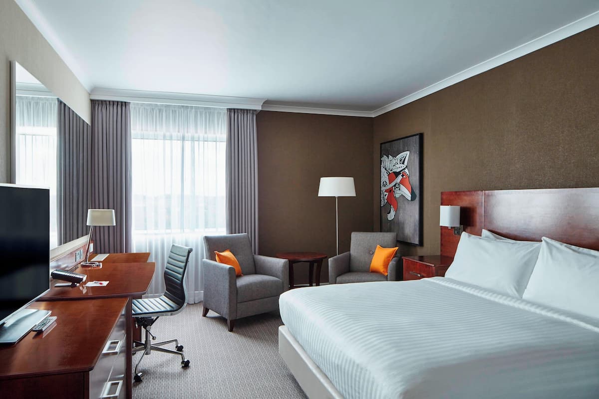 A hotel room with a large bed, two armchairs, a desk with a computer, a TV, and a painting on the wall.