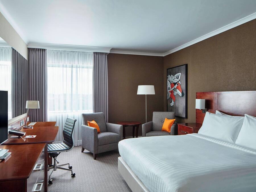 A hotel room with a large bed, two armchairs, a desk with a computer, a TV, and a painting on the wall.