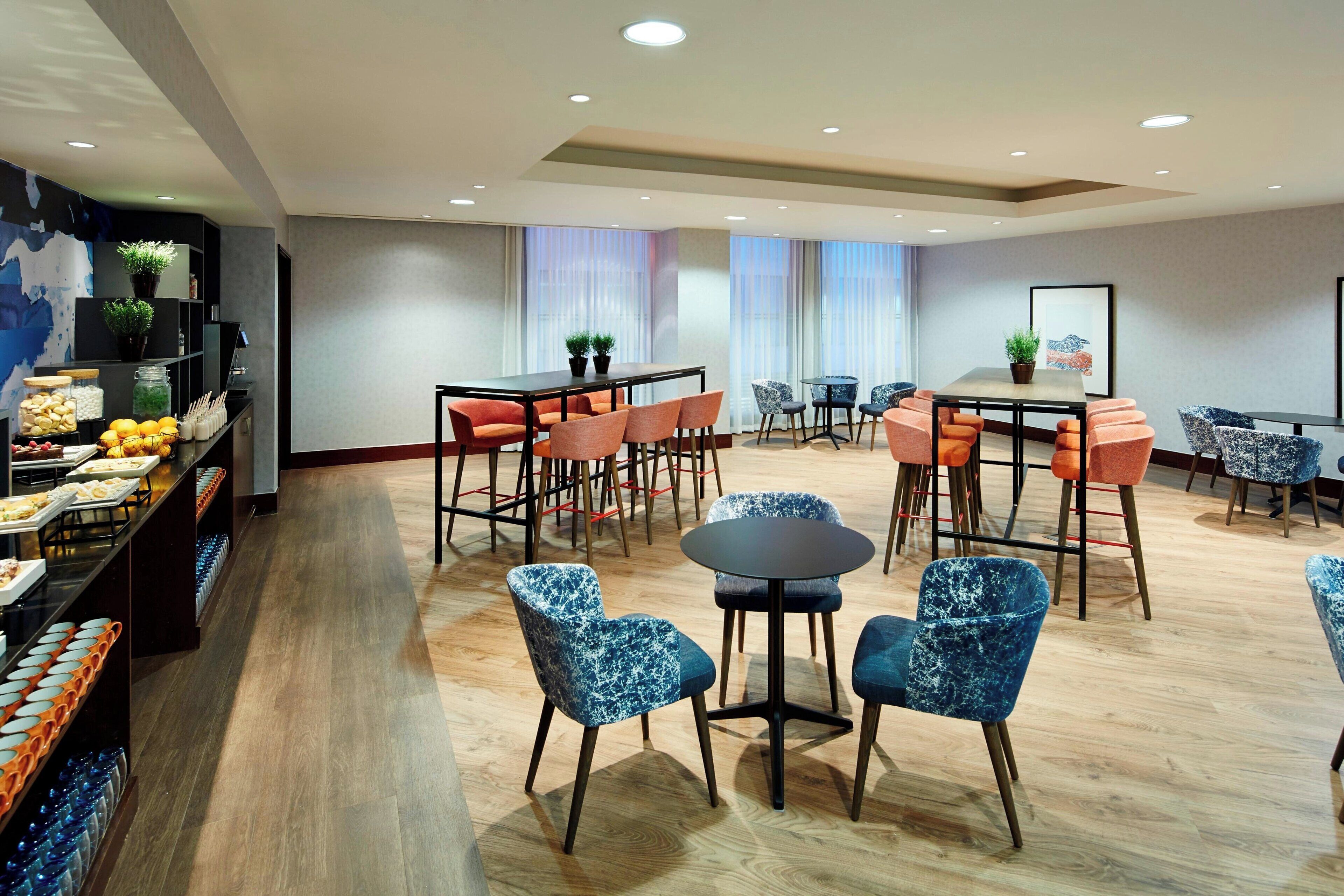 A modern dining area with round tables and blue patterned chairs, a buffet with fruits and vegetables, and potted plants.