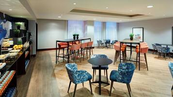 A modern dining area with round tables and blue patterned chairs, a buffet with fruits and vegetables, and potted plants.