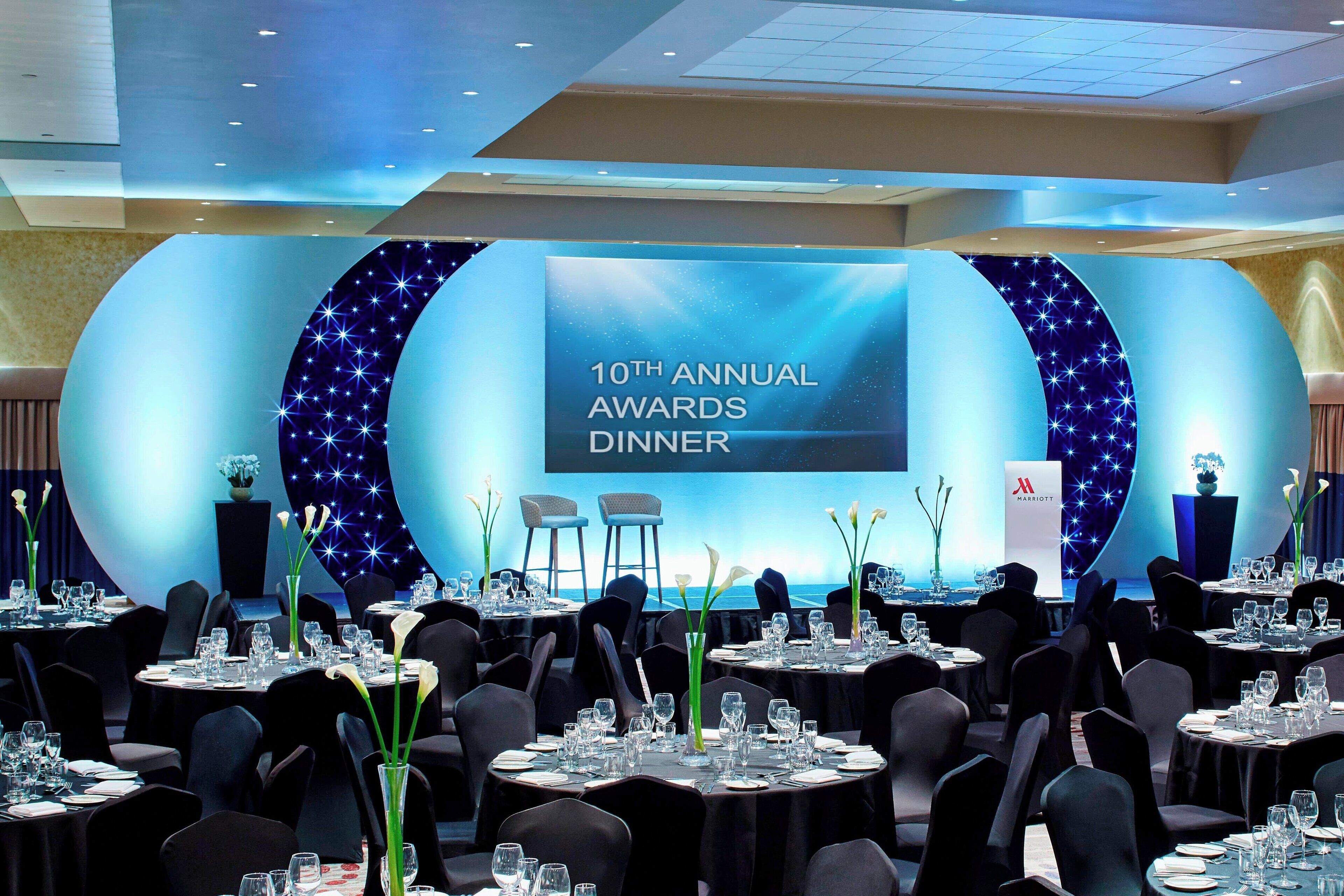 Un espacio para eventos formales con mesas dispuestas para una cena, un escenario con una pantalla que muestra '10ma Cena Anual de Premios', y vista al mar de fondo.