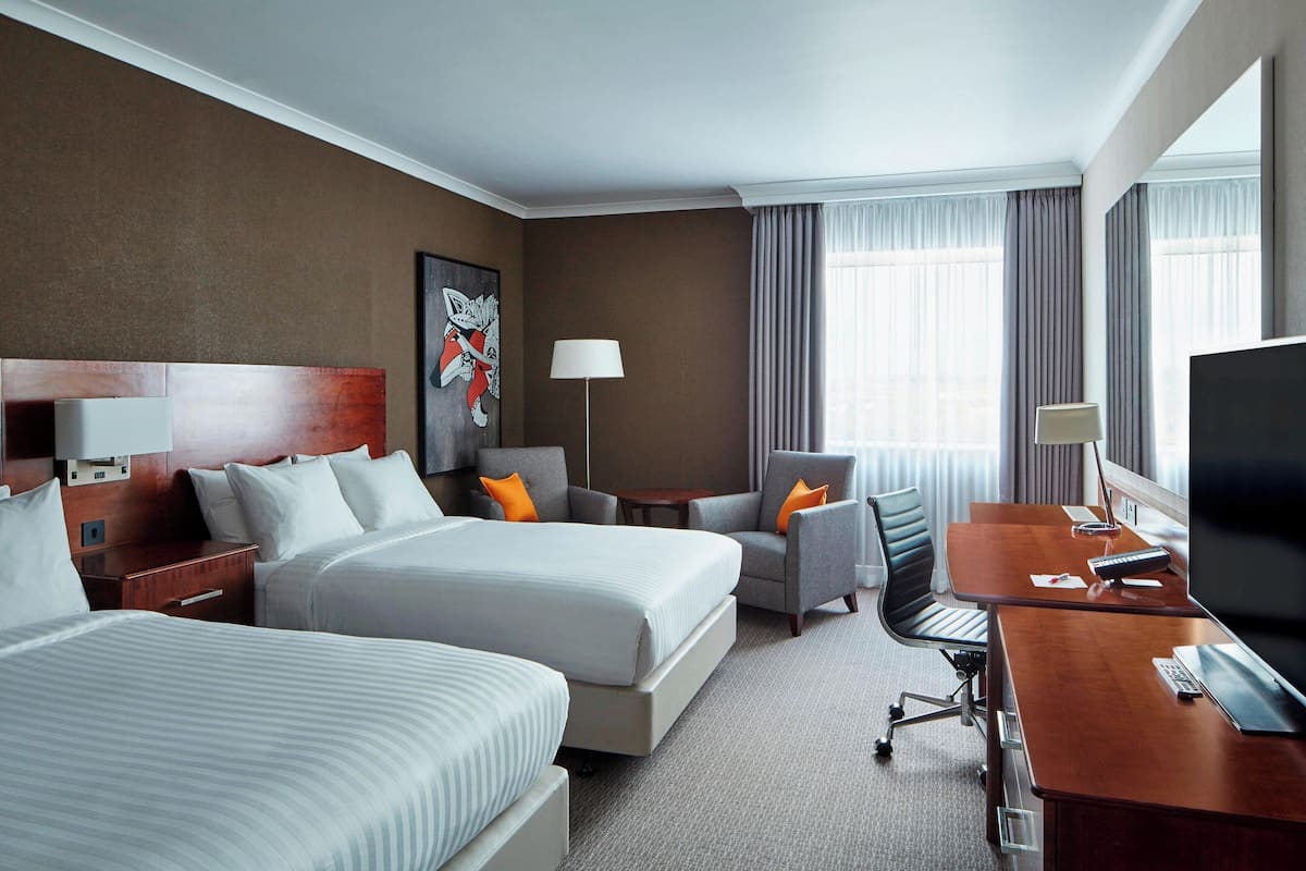 A hotel room with two beds, a desk, a chair, a TV, and a lamp.