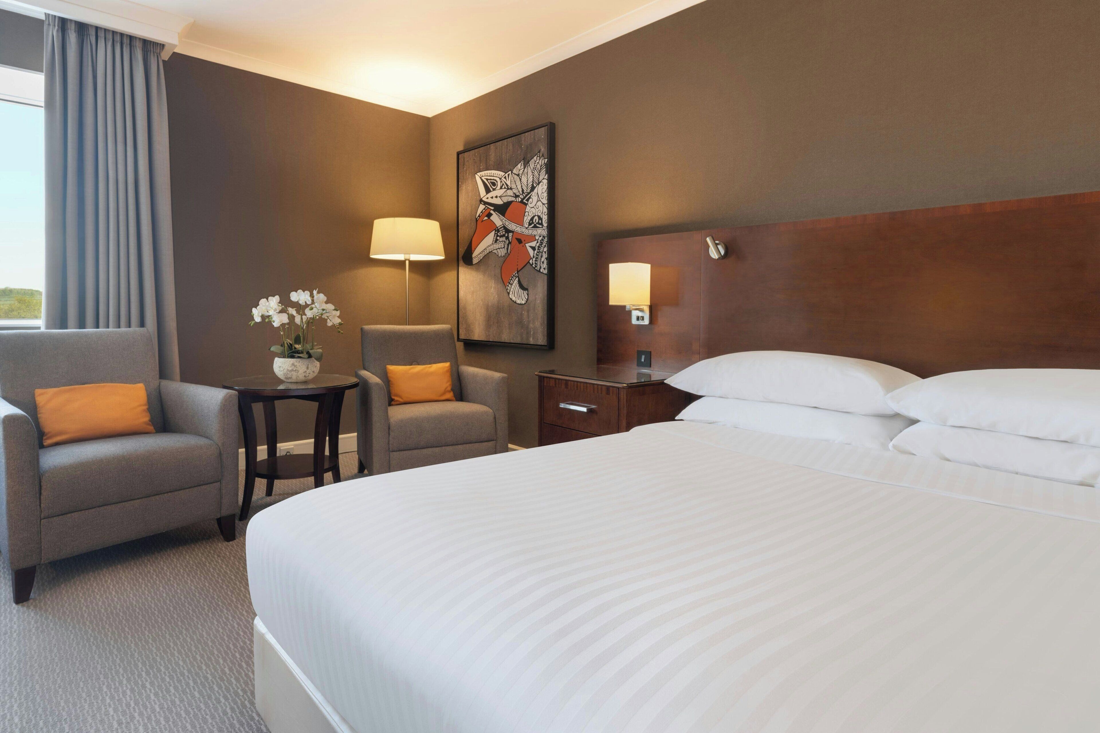 A hotel room with a large bed, two armchairs, a small table with a vase of flowers, a painting on the wall, and a nightstand with a lamp.