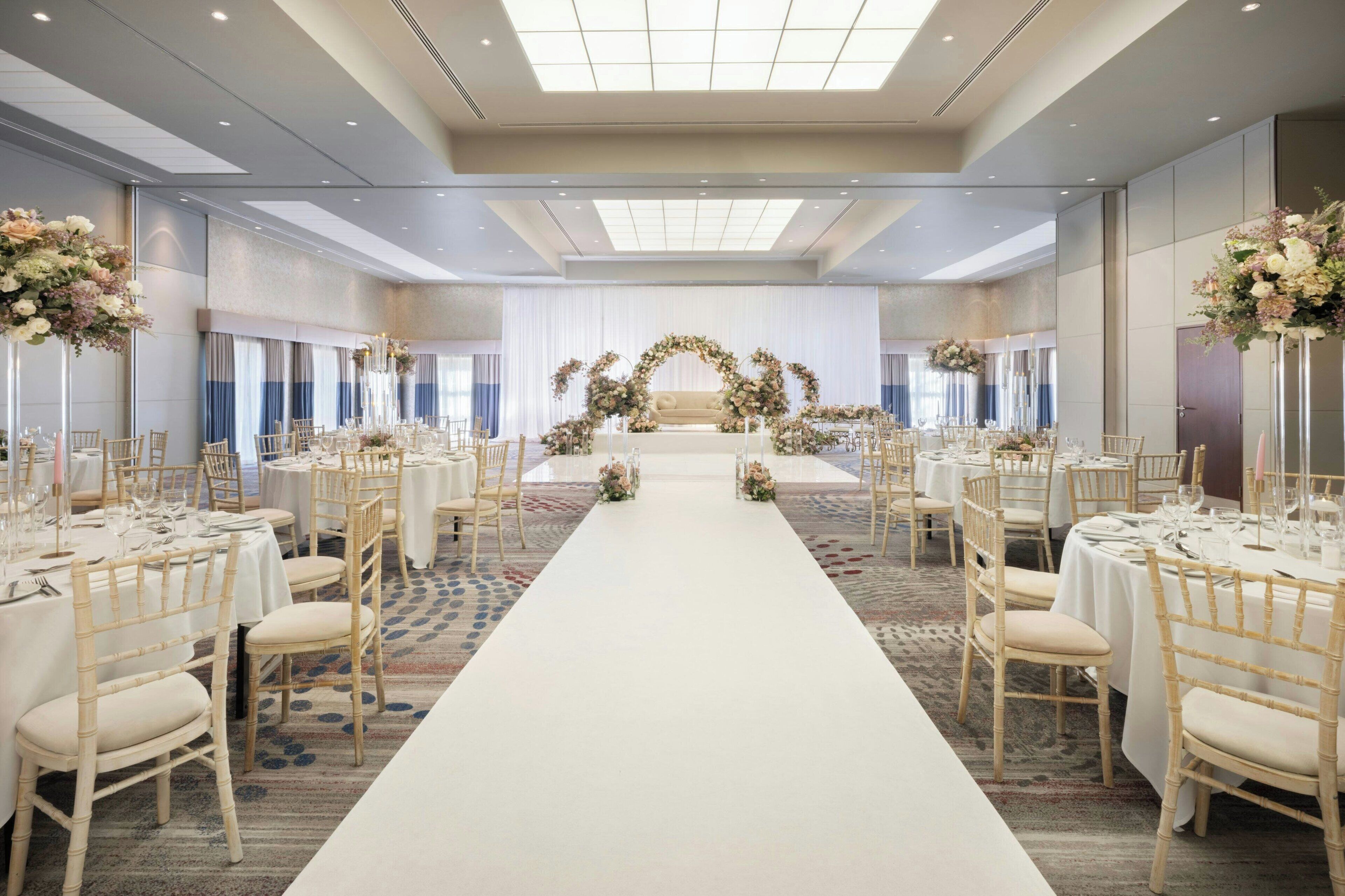 A spacious banquet hall with a central stage, rows of tables with chairs, and floral arrangements.