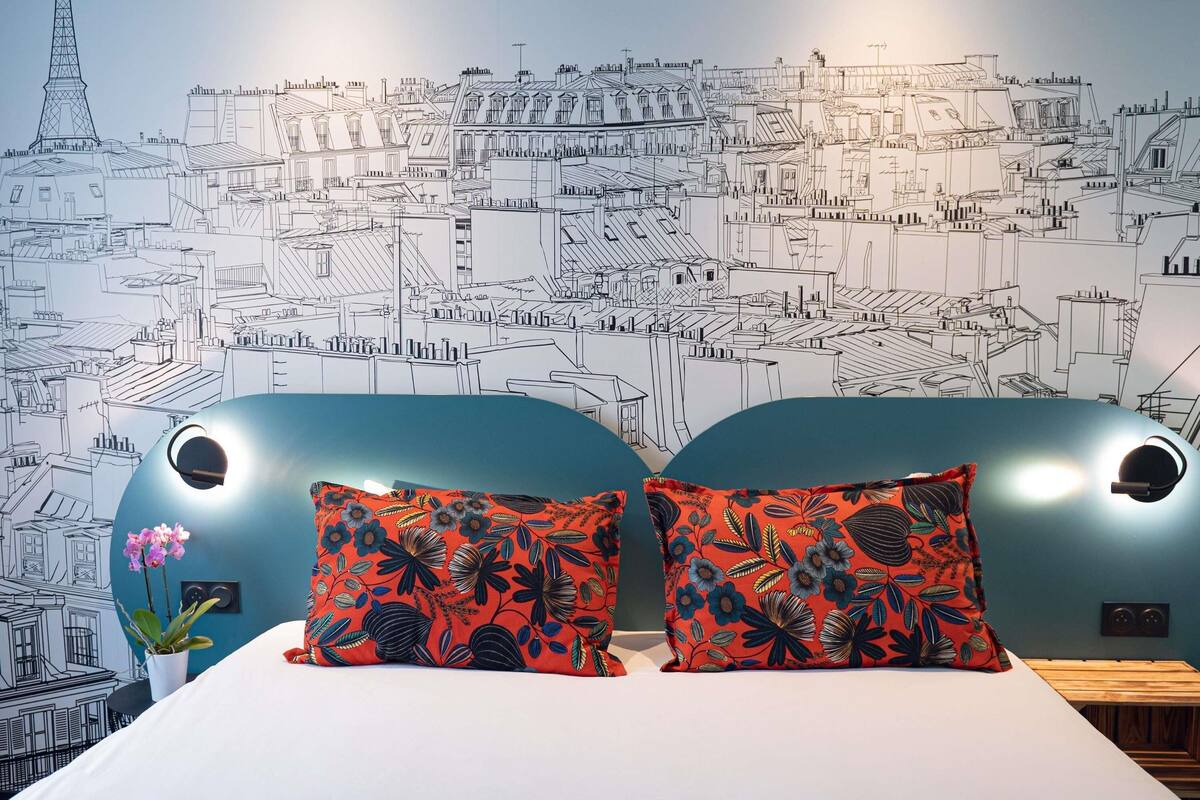 A hotel room with a large bed, two pillows with a floral pattern, a potted plant, and a wall mural of a cityscape.