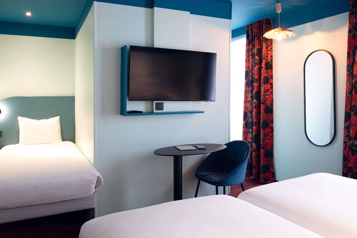 A hotel room with two beds, a flat-screen TV mounted on the wall, a small round table with a chair, and a large mirror.