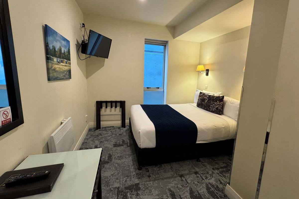 Ein Hotelzimmer mit einem Bett, einem Schreibtisch mit Fernbedienung, einem an der Wand befestigten Fernseher, einem Fenster und einem Nachttisch mit Lampe.