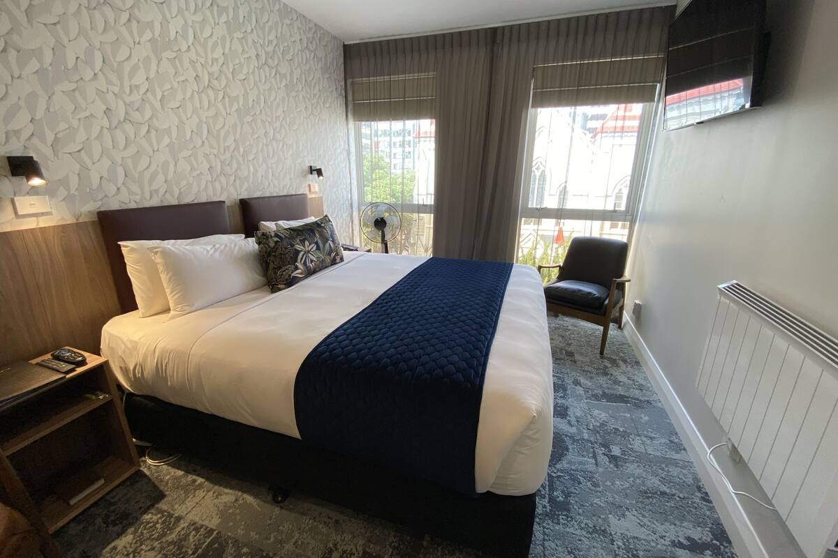 Ein Hotelzimmer mit einem großen Bett, einem Nachttisch, einem Sessel, einem an der Wand befestigten Fernseher und einem Fenster mit Vorhängen.