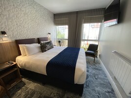 A hotel room with a large bed, a nightstand, a chair, a wall-mounted TV, and a window with curtains.