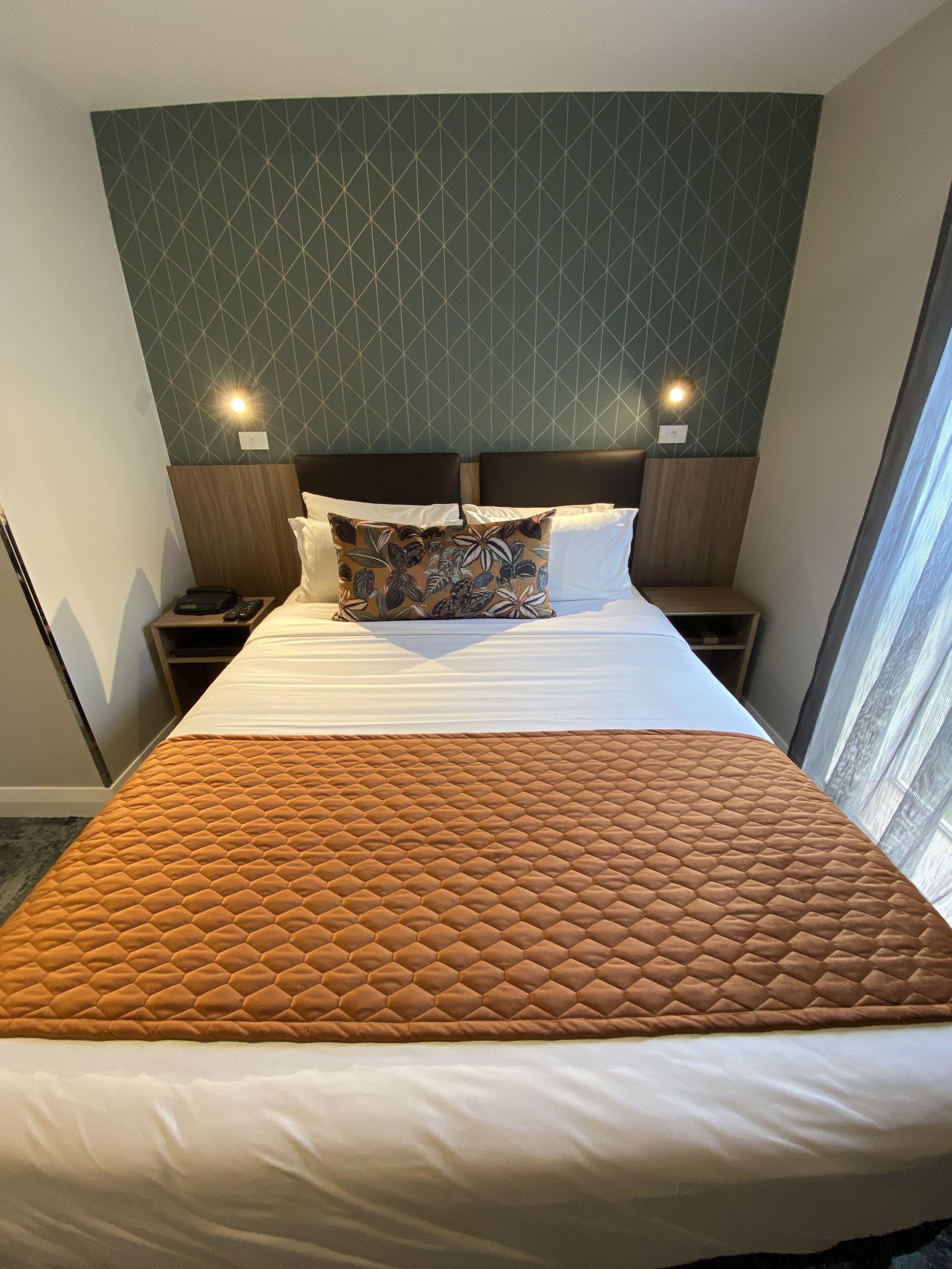Une chambre d’hôtel avec un grand lit, une couvre-lit orange matelassée, un coussin à motifs et un décor mural géométrique.