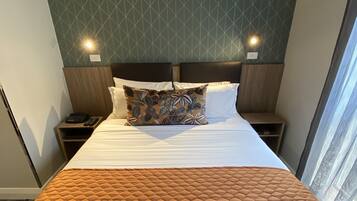 Une chambre d’hôtel avec un grand lit, une couvre-lit orange matelassée, un coussin à motifs et un décor mural géométrique.