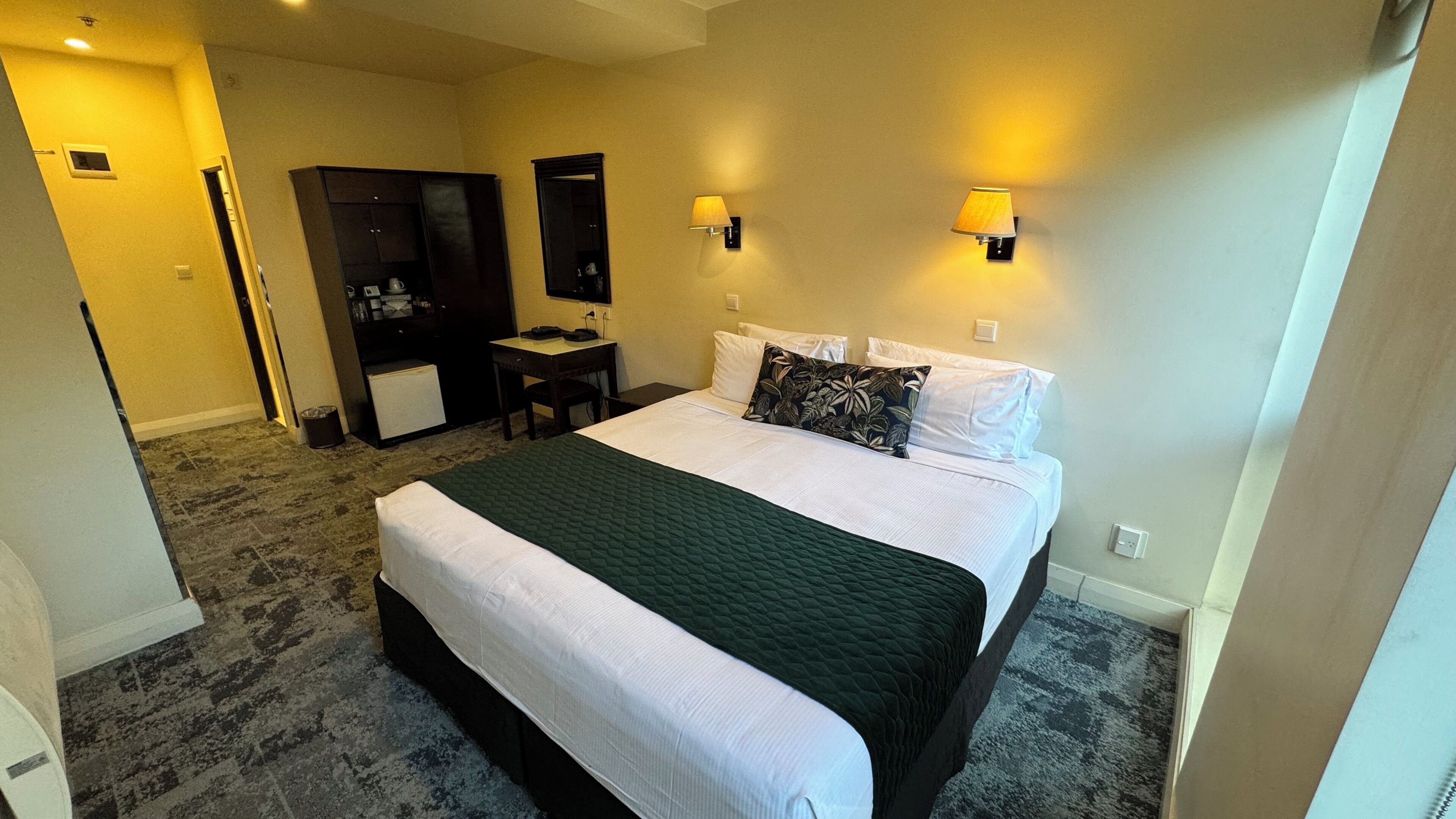 Habitación de hotel con cama, mesitas de noche, escritorio, armario y dos lámparas fijadas a la pared.