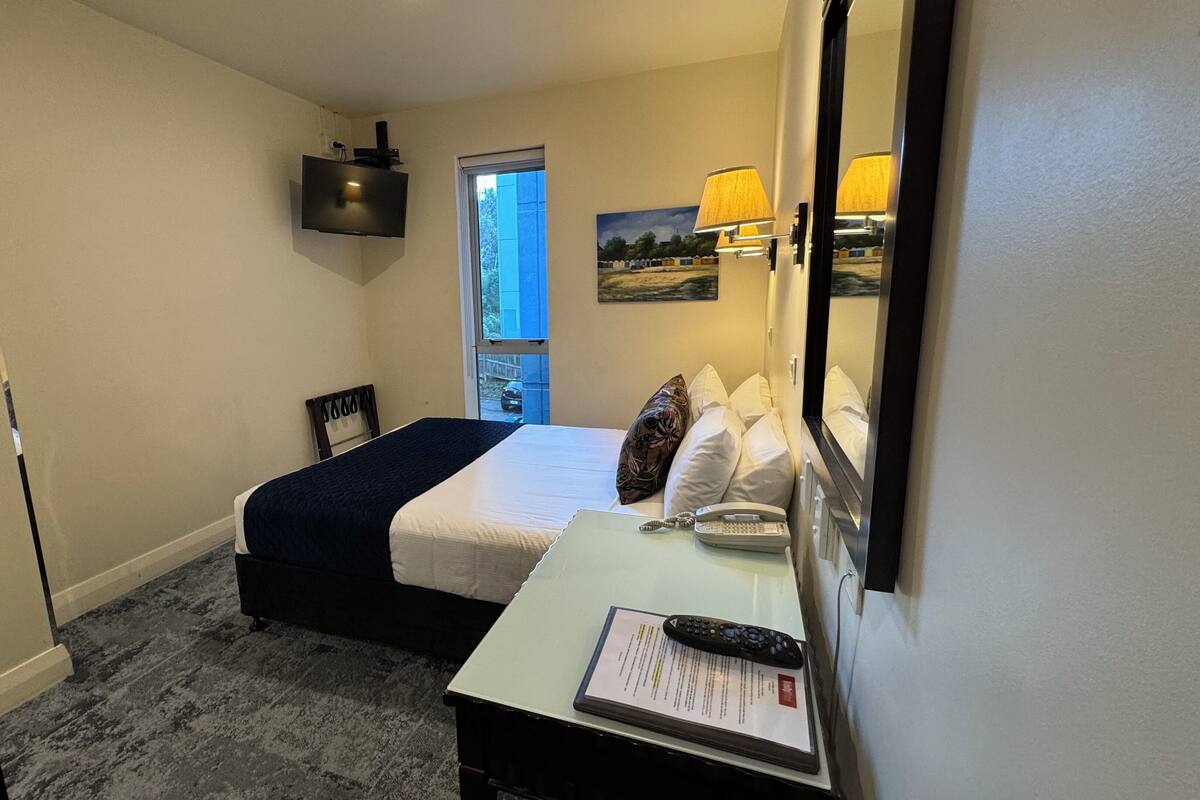 Ein Hotelzimmer mit einem Bett, einem Schreibtisch mit Telefon und Fernbedienung, einem Spiegel und einem Fenster mit Blick nach außen.