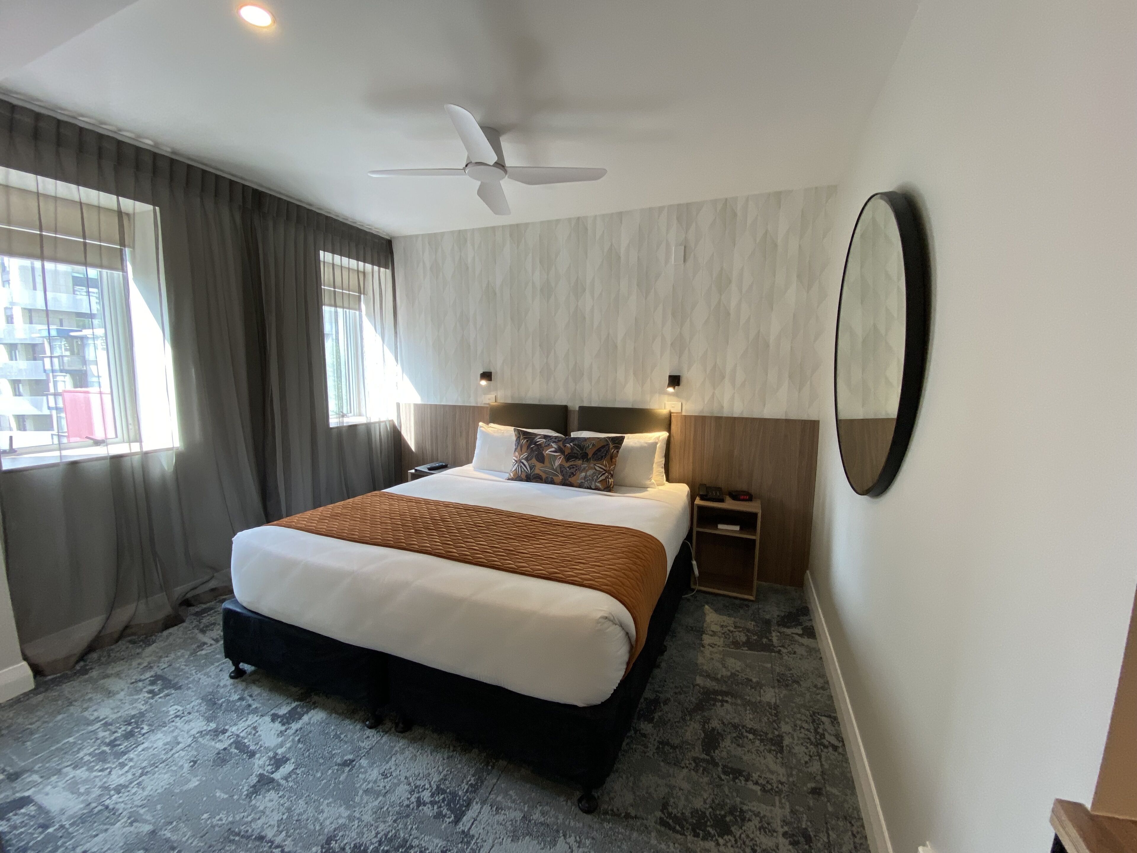 A hotel room with a large bed, a ceiling fan, a bedside table, a nightstand, and a large mirror.