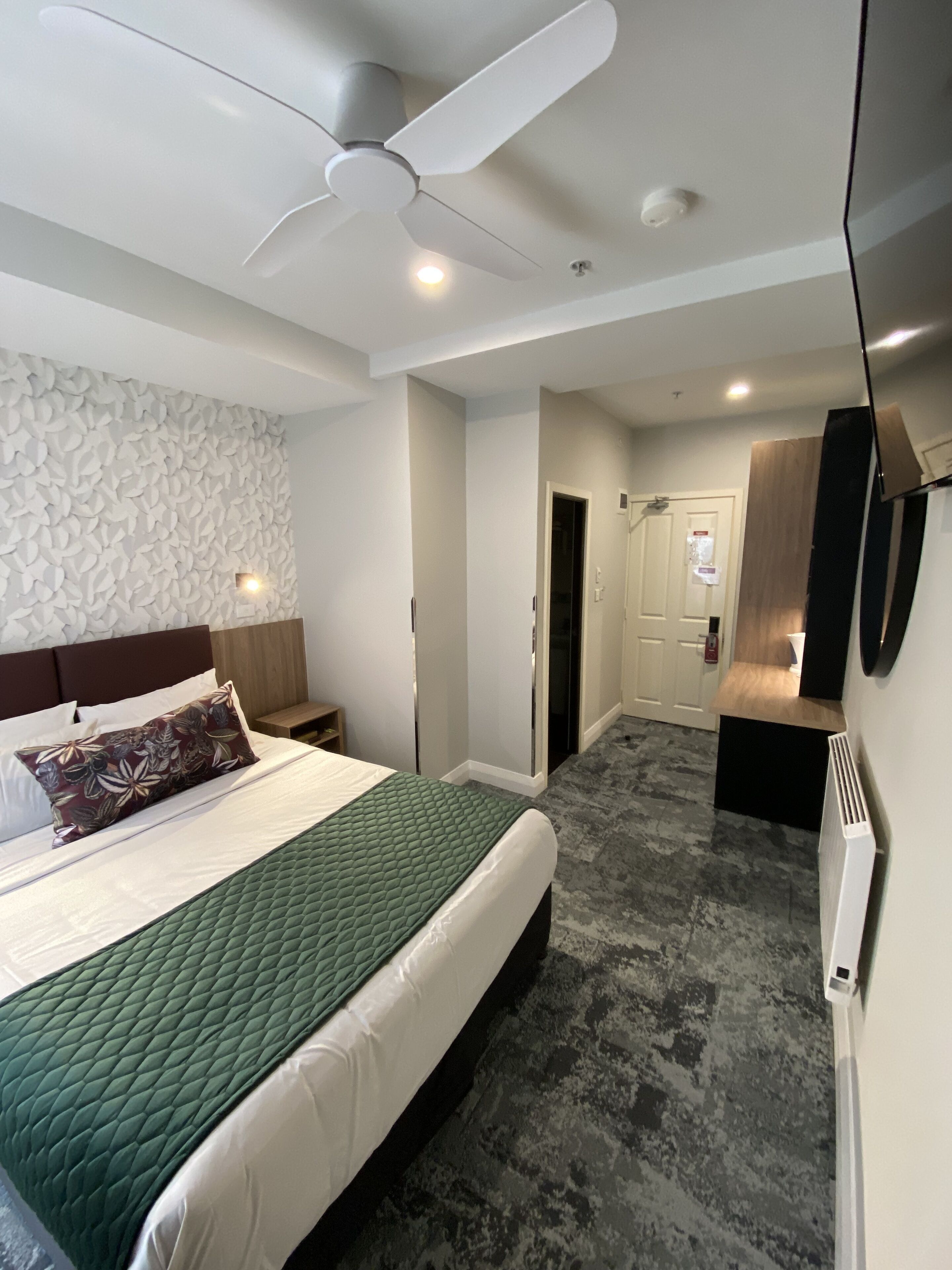 A hotel room with a bed, a ceiling fan, a wall-mounted TV, and a textured wall.