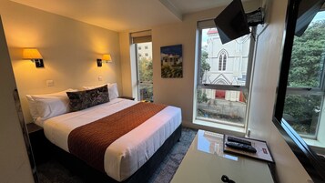 A hotel room with a bed, two wall-mounted lamps, a window with a view of a building, and a desk with a remote control.