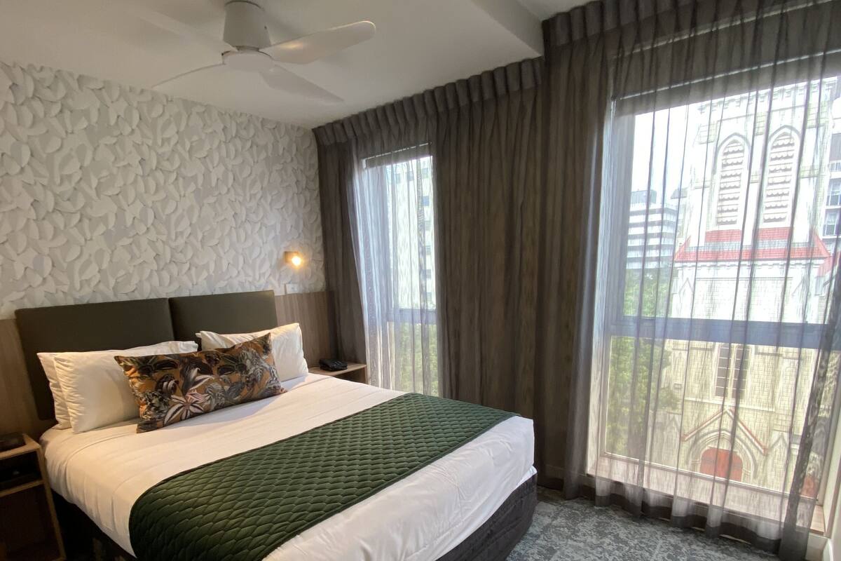 Ein modernes Hotelzimmer mit einem großen Bett, einem Deckenventilator und Blick auf ein Gebäude durch transparente Vorhänge.