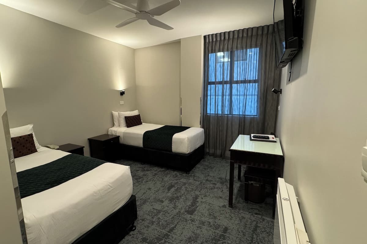 Ein Hotelzimmer mit zwei Betten, einem Schreibtisch, einem Sessel, einem Fernseher und einem Deckenventilator.