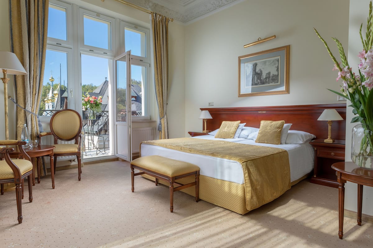 Quarto de hotel com uma cama grande, cabeceira de madeira, mesa de cabeceira com abajur, uma cadeira e vista para o exterior através de uma janela ampla.