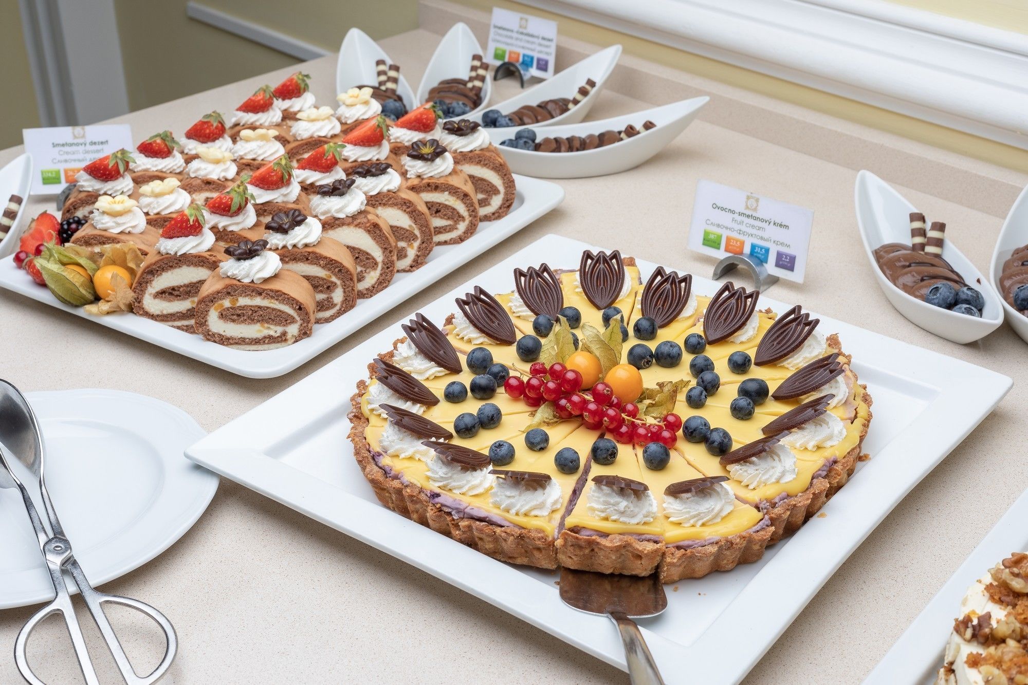 Diversos doces em exposição, incluindo uma tarte de chocolate com mirtilos e groselhas vermelhas, um pastel de creme com morangos e um rolo de chocolate recheado com creme.