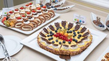 Diversos doces em exposição, incluindo uma tarte de chocolate com mirtilos e groselhas vermelhas, um pastel de creme com morangos e um rolo de chocolate recheado com creme.