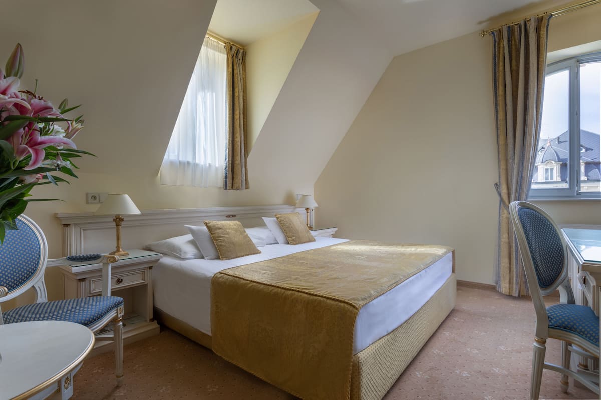 Quarto de hotel com cama, duas mesas de cabeceira, uma cadeira, escrivaninha e janela com cortinas.