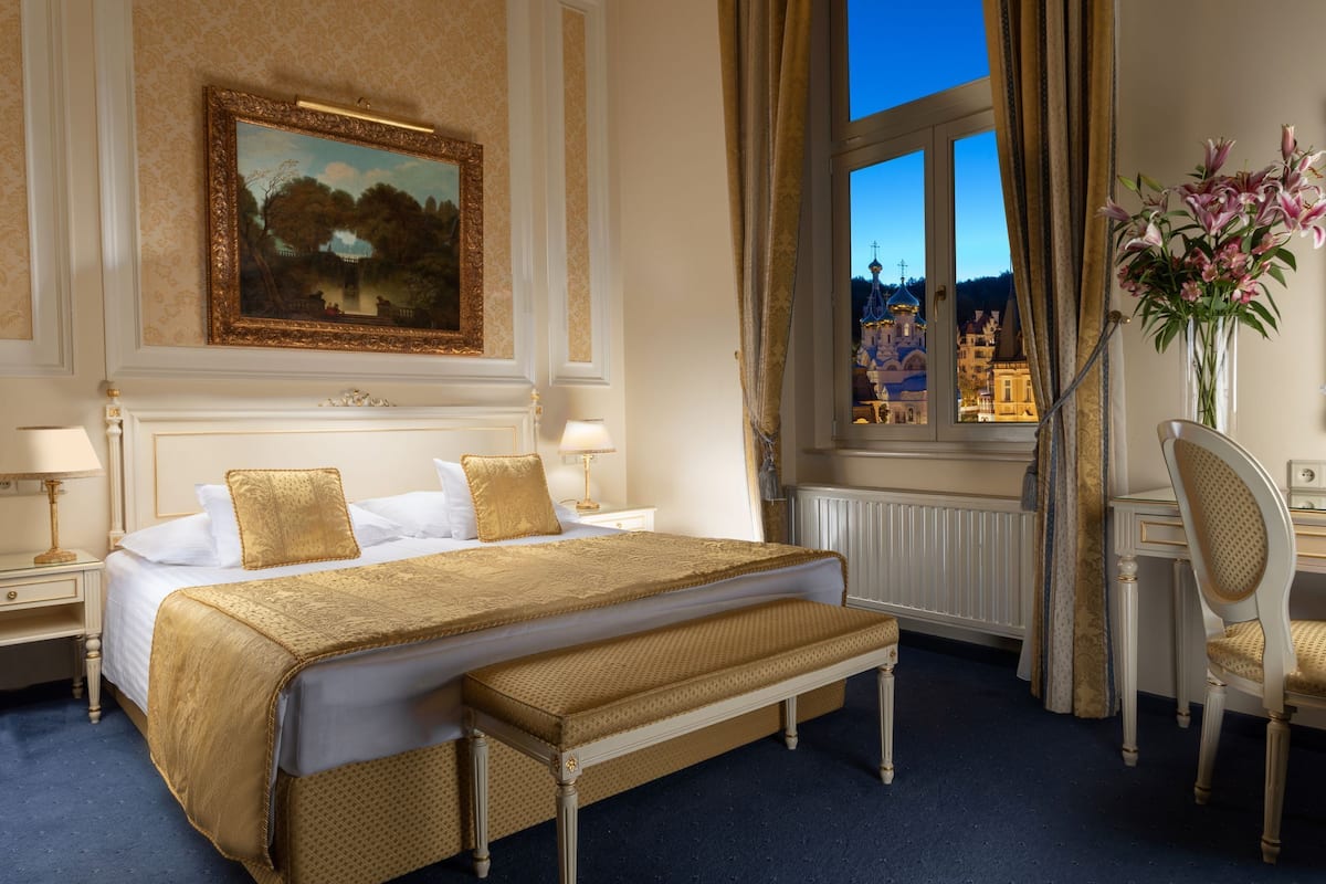 Quarto de hotel com uma cama grande, uma escrivaninha, uma cadeira, uma janela com vista para um edifício e um quadro na parede.